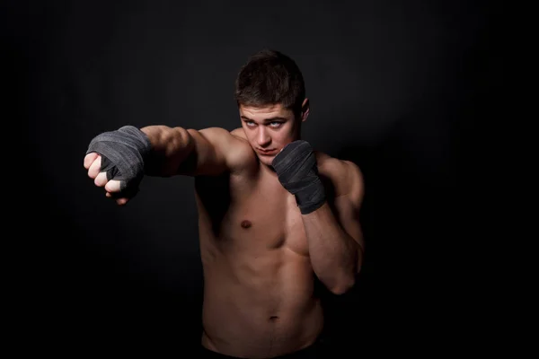 Мускулистый спортсмен боксирует на черном фоне. Мужчина с голым туловищем в шортах, руки перемотанные бинтами для бокса . — стоковое фото