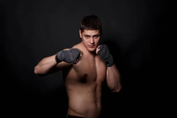 Hombre atlético muscular boxeando sobre un fondo negro. Un hombre con un torso desnudo en pantalones cortos, manos peremotanye vendajes para el boxeo . — Foto de Stock