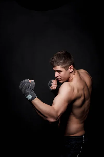 Uomo atletico muscolare boxe su uno sfondo nero. Un uomo con un busto nudo in pantaloncini, mani peremotanye bende per la boxe . — Foto Stock