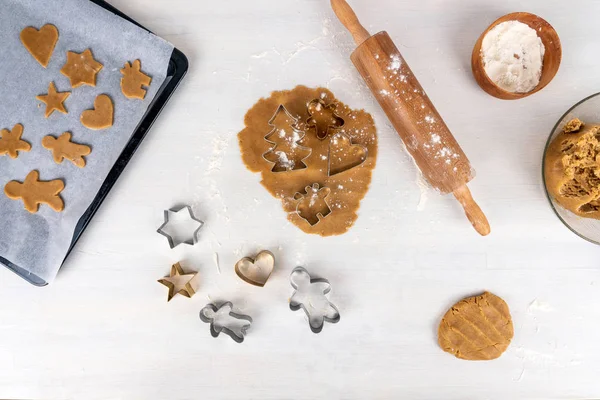 Gingerbread baking concept with gingerbread dough, flour, rolling pin and gingerbread cookies on baking tray.  Copy space for text on white kitchen table.