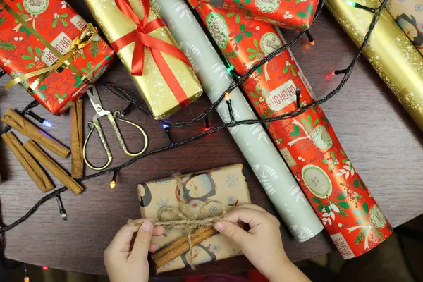 小さな女の子はテーブルの上に着色された紙でクリスマスプレゼントをパックします。家族の伝統. — ストック写真