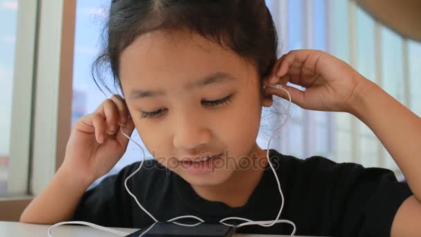 Asiática niña usando teléfono inteligente con auriculares en el oído y sentado cerca de ventanas de vidrio — Vídeos de Stock