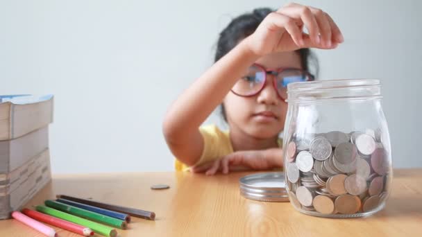 Asiática niña poner la moneda en un frasco de vidrio transparente en la metáfora de la mesa ahorro de dinero concepto con sonido seleccionar el enfoque en el frasco — Vídeo de stock