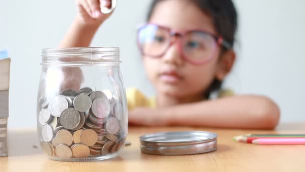 Asian little girl putting the coin into a clear glass jar on table metaphor saving money concept with sound select focus on jar — Stock Video