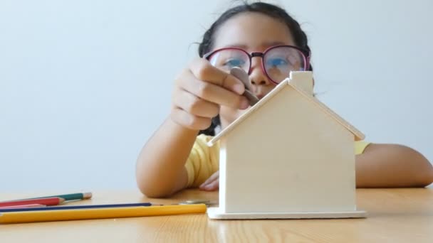 Asiatico bambina mettendo soldi moneta in chiara casa banca metafora risparmio di denaro per comprare la casa e il concetto immobiliare con suono ambientale — Video Stock