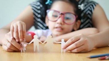 Yakın çekim Asyalı küçük kız Tay anaokulu öğrenci üniforma ve kesim beyaz kağıt yapma aile şekil baba anne oğul ve odak alan sığ derinliği seçin kızı ahşap masa üzerinde oynarken annesi