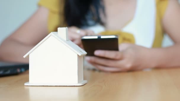 Mãos de mulher usando o computador de telefone inteligente e colocando moeda de dinheiro na metáfora da casa do banco porquinho cálculo de aplicativo móvel para economizar dinheiro para comprar casa e conceito imobiliário selecione o foco na casa — Vídeo de Stock