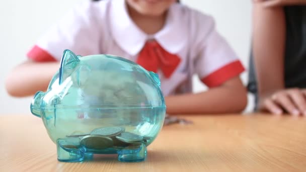 Asian little girl in Thai kindergarten student uniform putting money coin into clear piggy bank with hands of mother select focus on pig saving money for education concept with ambient sound — Stock Video