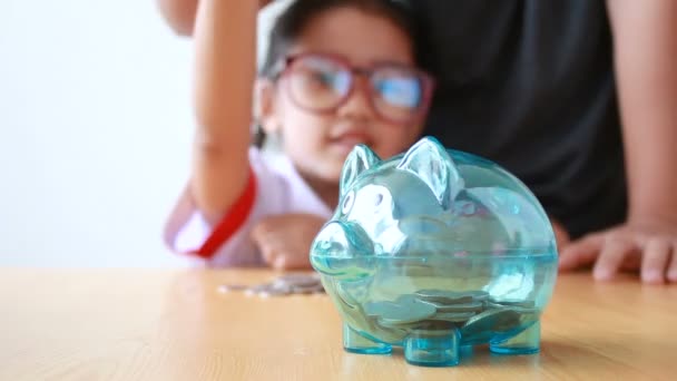 Asiática niña en tailandés kindergarten estudiante uniforme poner dinero moneda en claro alcancía con las manos de la madre seleccionar se centran en cerdo ahorrar dinero para la educación concepto con ambiente sonido — Vídeos de Stock