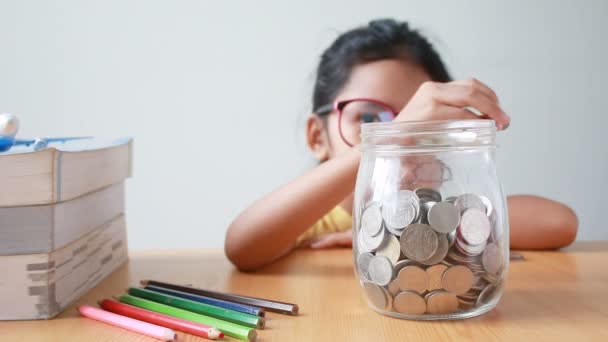 Kitap eğitim kavramı için para tasarrufu tablo metafor üzerinde bir açık cam kavanoza para koyarak Asyalı küçük kız — Stok video