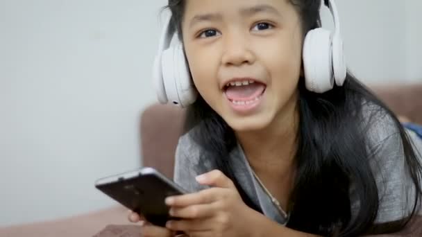 Asyalı küçük kız mutluluk konuşma başparmak ile beyaz kablosuz kulaklık ile müzik dinlemek ve kameraya bak — Stok video
