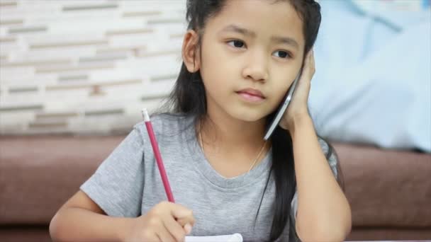 Закройте снимок маленькой азиатской девочки карандашом, делающей домашнюю работу, и поговорите со смартфоном — стоковое видео