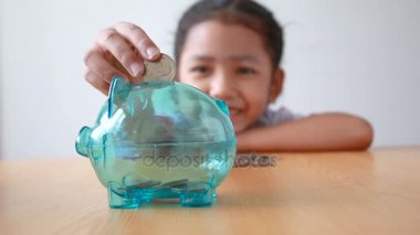 Asyalı küçük kız Tay anaokulu öğrencisi para sikke içine koyarak Tekdüzen temizleyin kumbara ahşap masa mecaz para tasarrufu için eğitim kavramı üzerinde