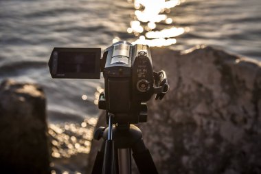 Bir tripodun üzerindeki kamera denizin üzerinde gün batımını çekiyor.