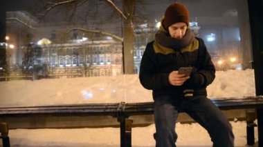 bir akıllı telefon kullanarak gri ve bulutlu günde bir otobüs durağında oturan bir adam Close-Up