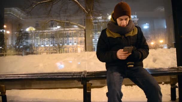 Крупним планом чоловік, який сидить на зупинці в сірий і хмарний день, використовуючи смартфон — стокове відео