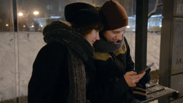 Giovane uomo e donna che utilizzano touch pad sulla stazione degli autobus della città di notte, display a led informativi e persone che salgono sull'autobus in background — Video Stock