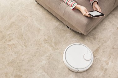 Kadın kanepede dinlenirken robot elektrikli süpürge odayı temizliyor.