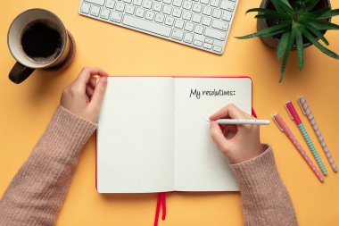 Stock photo of a young woman hands writing in a 2020 new year notebook with list of resolutions and objects on yellow background clipart