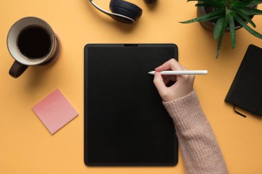 Sarı arka planda dijital tablete yazı yazan bir kadının el resmi.