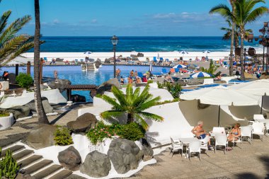 Puerto de la Cruz, Tenerife, İspanya - 18 Aralık 2019: İspanyol güneşinin tadını çıkaran turistler, Cesar Manrique tarafından tasarlanan Puerto de la Cruz 'da bulunan açık hava yüzme havuzu kompleksi.