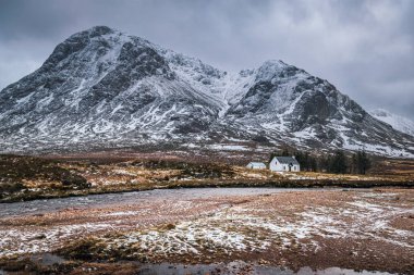 Glen Coe 'nun dramatik kış manzarası. Buachaille' in eteklerinde ikonik beyaz kır evi. Etive Mor, River Coupall ve ufukta fırtınalı bulutlar..