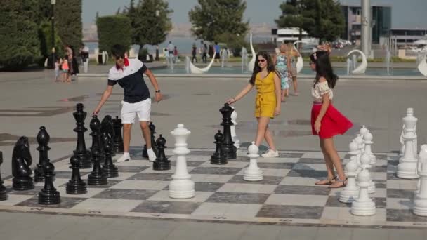 Plaza de la ciudad con enormes piezas de ajedrez. Adolescentes en ropa colorida juegan al ajedrez — Vídeo de stock