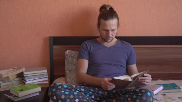 Jovem bonitão está deitado na cama e lendo. Muito tempo livre, muitos livros — Vídeo de Stock