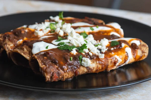 Vegetarian Mexican food, enfrijoladas. A type of enchilada dish of fresh cheese wrapped in corn tortilla and bathed in a spicy black bean sauce, topped with cream, cheese, spicy sauce and cilantro — Stock Image Vegetarian Mexican Food Enfrijoladas Type Enchilada Dish Fresh Cheese Wrapped Royalty Free Stock Photos