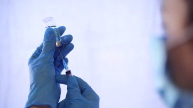 Tıbbi maskeli ve mavi eldivenli doktor cam şişeden şırınga dolduruyor. Doktorun elleri yakın plan. Hastane ve ilaç