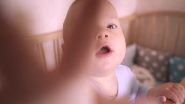 ベッドの中で面白い赤ちゃんが立ってカメラを見ています 子供は手をカメラに伸ばします 子供部屋の子供はベビーベッドにいる 子供はカメラに手を伸ばしてそれを調べる — ストック動画