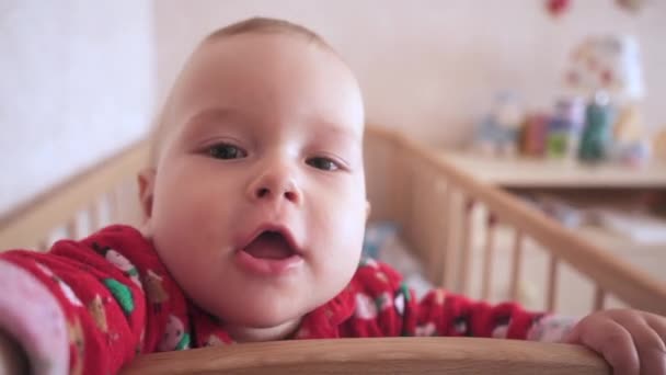 ベッドの中で面白い赤ちゃんが立ってカメラを見ています 子供は手をカメラに伸ばします 子供部屋の子供はベビーベッドにいる 子供はカメラに手を伸ばしてそれを調べる — ストック動画