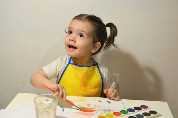 Uma criança pequena está sentada à mesa desenhando e sorrindo — Fotografia de Stock