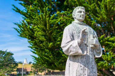 Monterey Presidio 'da Aziz Peder Junipero Serra heykeli. Arka plan: Bulanık John Drake Sloat anıtı ve yeşil selvi ağacı - Monterey, California, Usa - 2019
