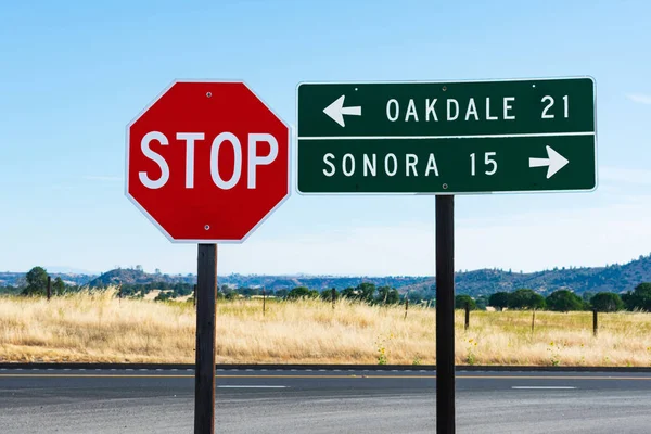 白い文字で8面の赤い停止記号と長方形の緑のガイド道路標識。オークデールとソノラへの方向と距離。背景カリフォルニアの象徴的な黄金の草と風景 — ストック写真
