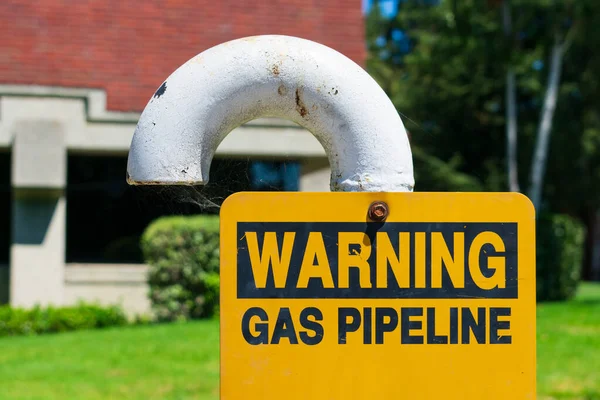 Gas pipeline warning sign on white bended pipe with blurred background of green landscape and commercial office building — 스톡 사진