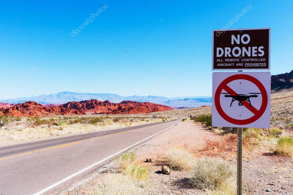 No drones zone warning sign prohibit the launching, landing, or operation of unmanned aircraft. Lonely road, scenic red sandstone formations, and desert scrub landscape background. Blue sky.