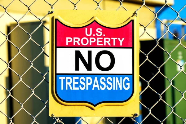 U.S. Property No Trespassing sign attached to a chain link fence with blurred industrial background