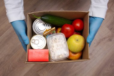Koruyucu tıp eldivenleri giymiş bir gönüllü, salgın sırasında ihtiyacı olanlar için yiyecek seti taşıyan bir kutu tutuyor. Temel gıdalar sebze gevreği makarna.