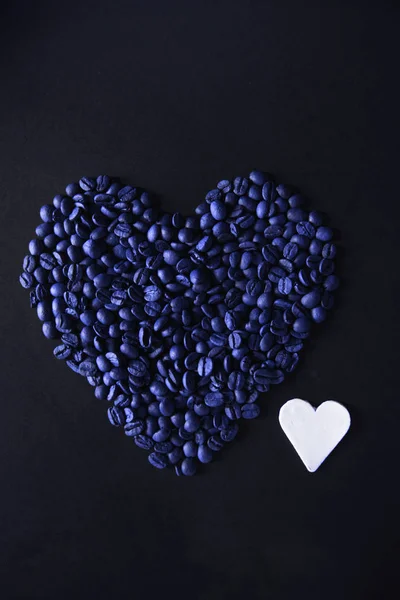 Corazón azul hecho de granos de café sobre un fondo negro con un corazón de arcilla blanca . —  Fotos de Stock
