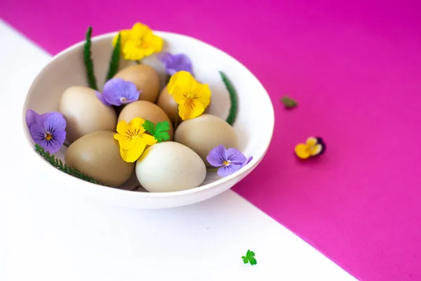 Пасхальные яйца с желтыми и сиреневыми цветами в белой миске на белом и розовом фоне. — стоковое фото
