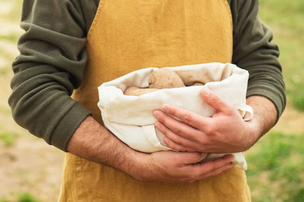 farmer in a working apron holds a bag of dug potatoes in his hands.