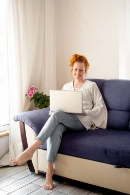 Güzel, mutlu bir genç kız, mavi bir kanepede oturup dizüstü bilgisayarını kullanıyor.