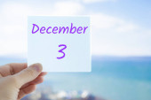 3. prosince. Ruka drží samolepku s textem 3. prosince na rozmazaném pozadí moře a oblohy. Kopírovat prostor pro text. Měsíc v koncepci kalendáře