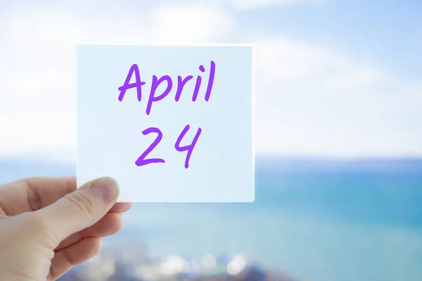 24. dubna. Ruka drží samolepku s textem 24. dubna na rozmazaném pozadí moře a oblohy. Kopírovat prostor pro text. Jarní měsíc v konceptu kalendáře — Stock fotografie
