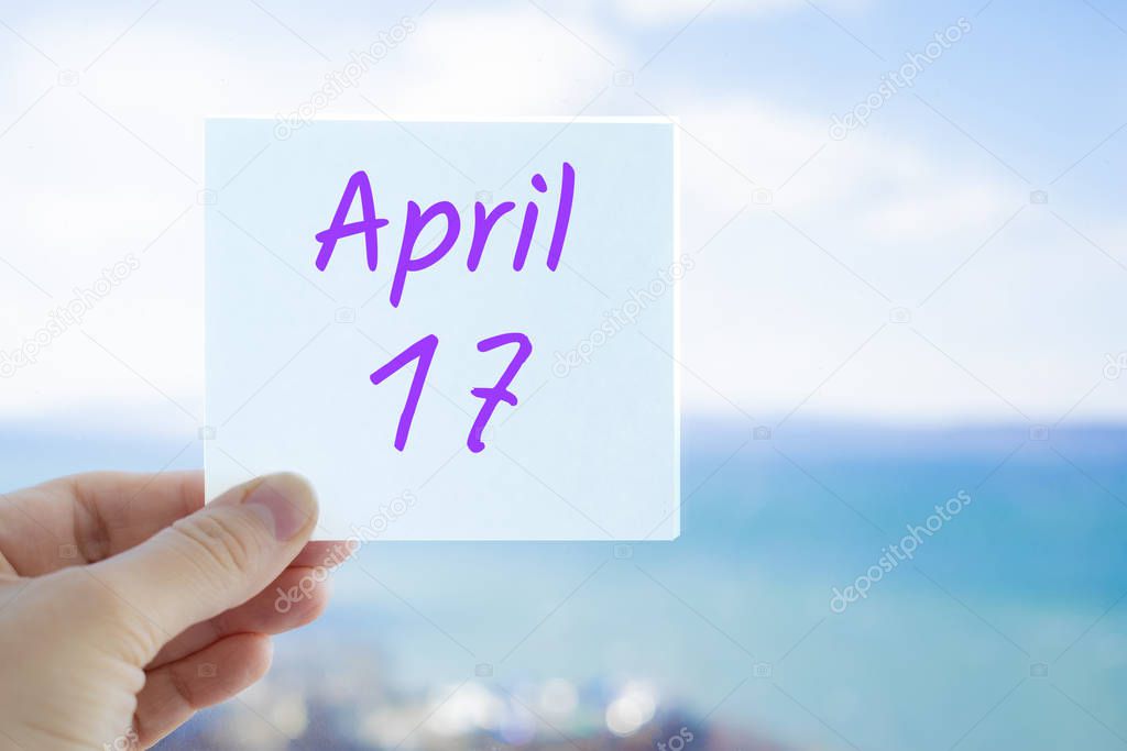 April 17th. Hand holding sticker with text April 17 on the blurred background of the sea and sky. Copy space for text. Spring month in calendar concept