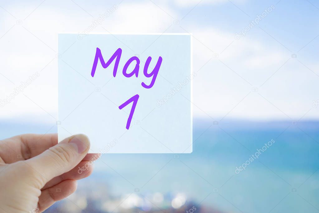 May 1st. Labour day and the world. Hand holding sticker with text May 1 on the blurred background of the sea and sky. Copy space for text. Month in calendar concept