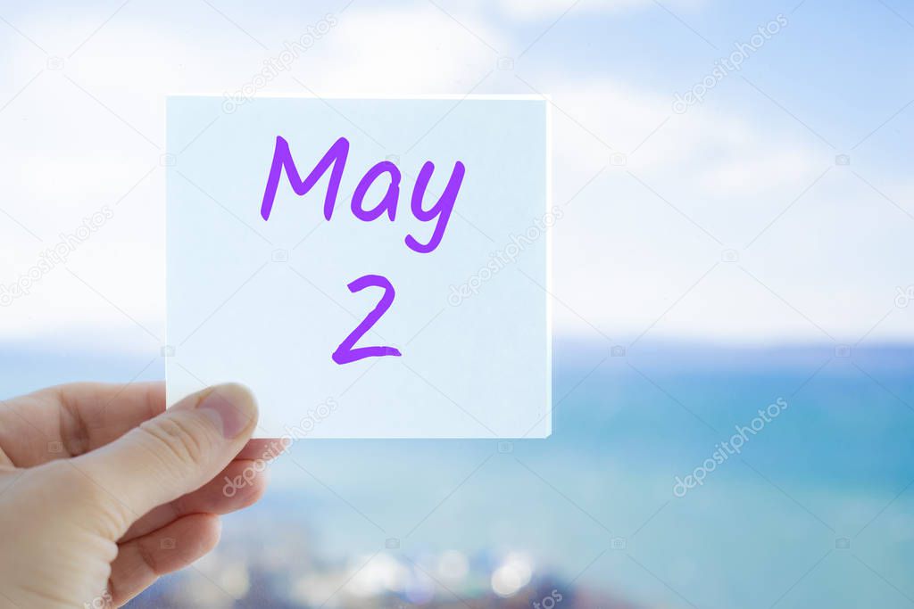 May 2nd. Hand holding sticker with text May 2 on the blurred background of the sea and sky. Copy space for text. Month in calendar concept
