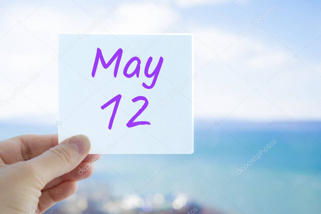 May 12th. Hand holding sticker with text May 12 on the blurred background of the sea and sky. Copy space for text. Month in calendar concept