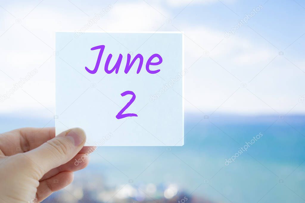 June 2nd. Hand holding sticker with text June 2 on the blurred background of the sea and sky. Copy space for text. Month in calendar concept